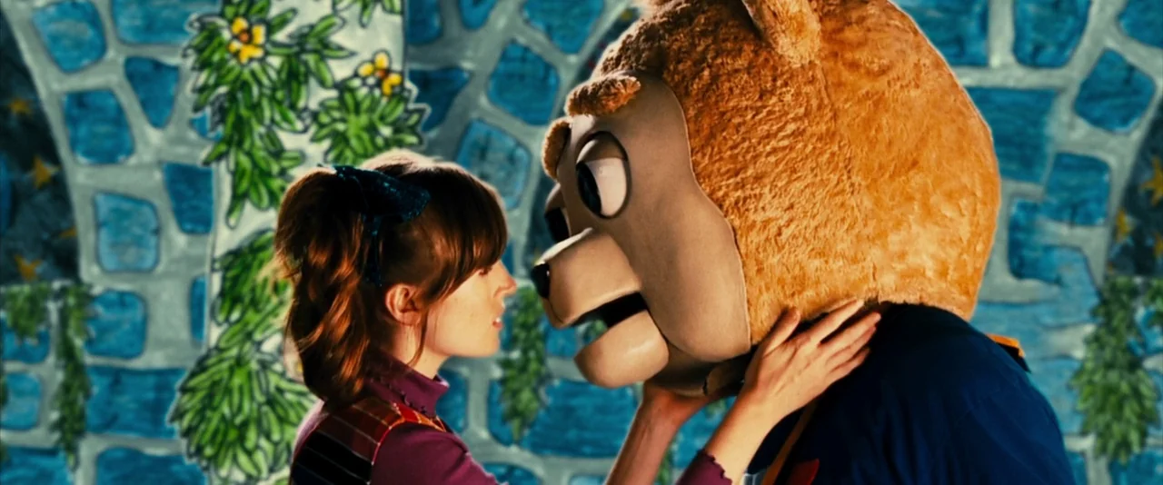 Brigsby Bear (2017) movie still: medium — A theatrical backdrop features painted blue stonework with hanging green plant illustrations. A woman…