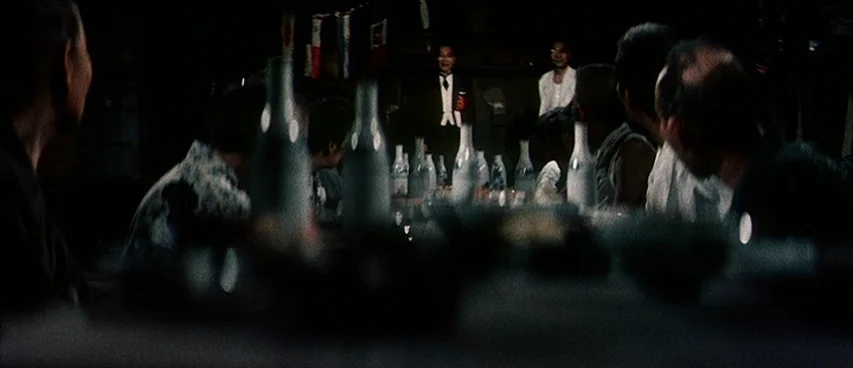 Jigoku (1960) movie still: medium — A dark, dimly lit room contains a group of men sitting at a table. A man in a suit stands at the far end of the…