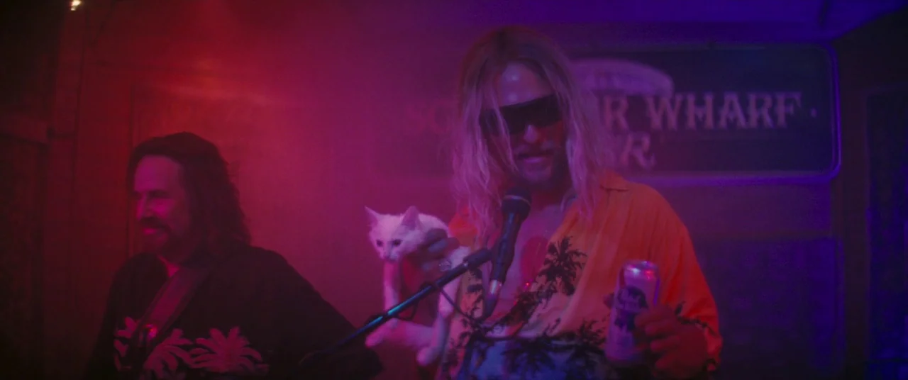 The Beach Bum (2019) movie still: medium — A dimly lit bar space is filled with purple and magenta haze. Two men stand on a small stage while one…