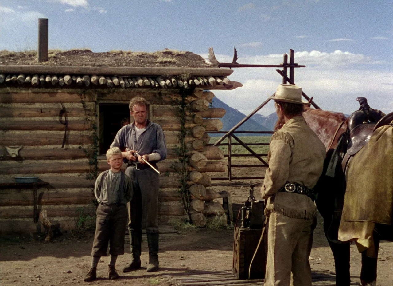 Shane (1953) movie still: fullbody — A rustic log cabin sits on a dirt lot with a wooden fence structure nearby. A man and young boy stand together…