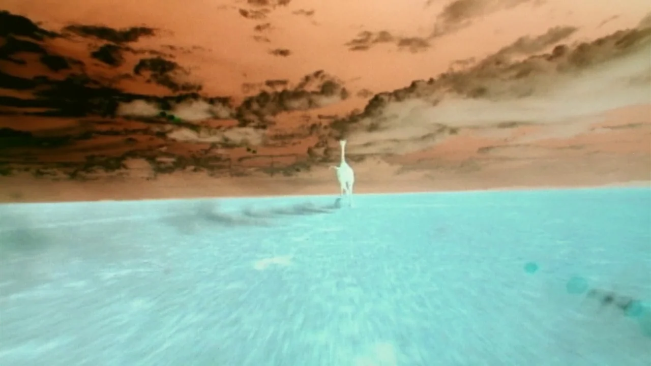 Naqoyquatsi (2002) movie still: wide — A giraffe stands alone in a wide, flat, and pale barren landscape. Dark clouds hang in the orange-toned sky…