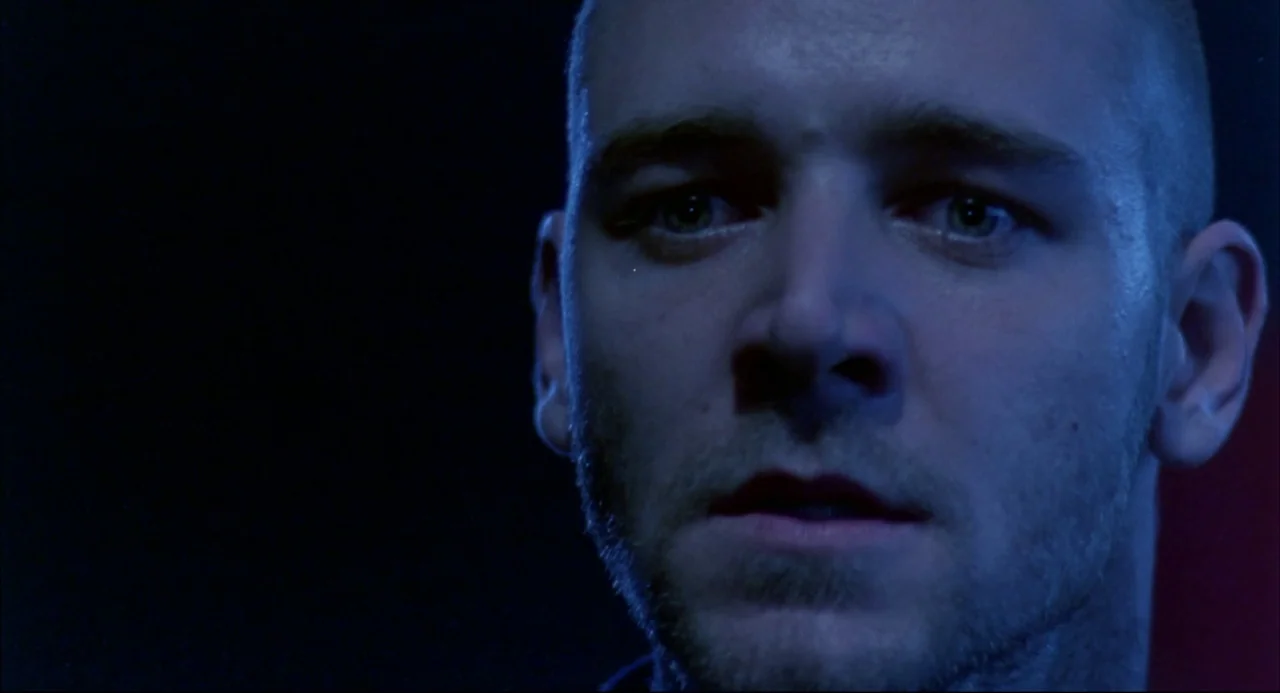 Romper Stomper (1992) movie still: closeup — A man's face is shown against a completely dark background. The subject stares ahead while bathed in…