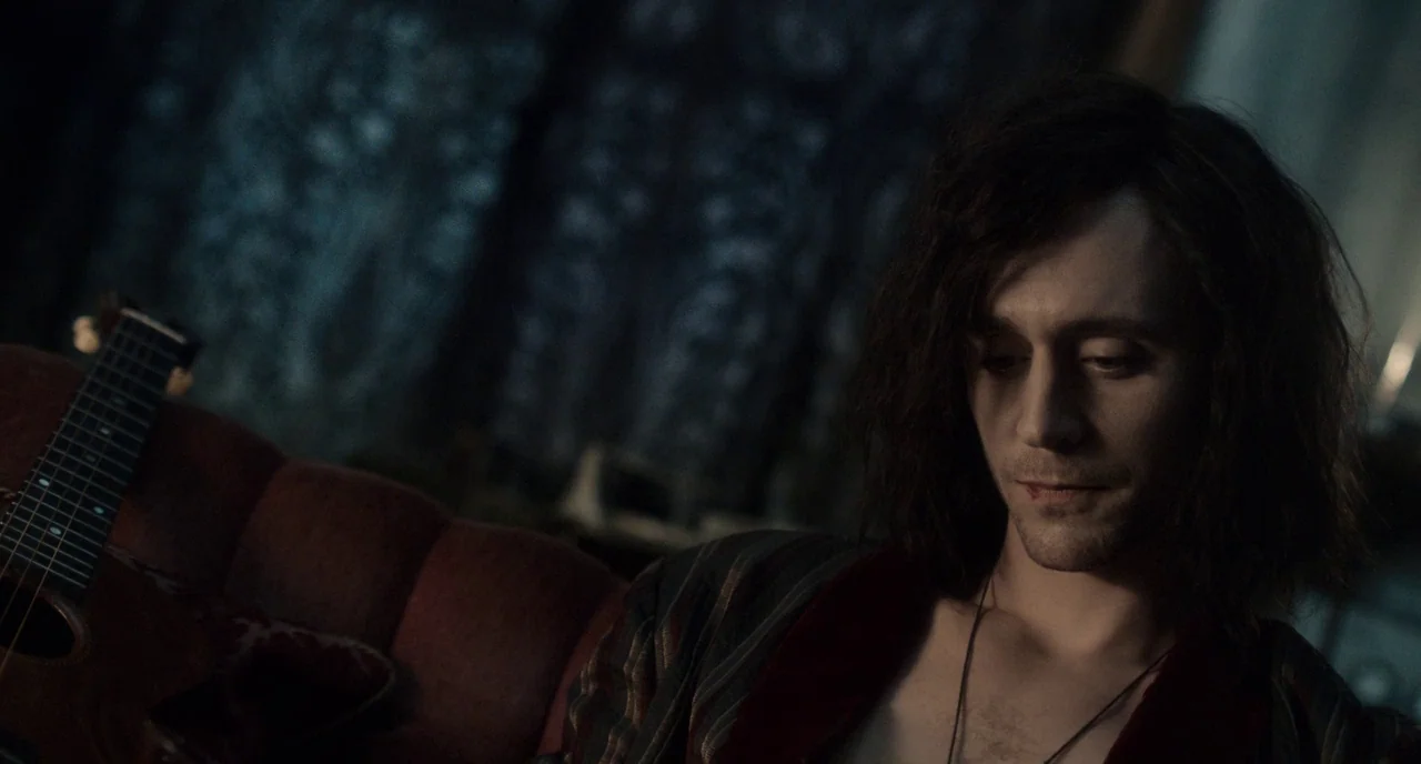 Only Lovers Left Alive (2013) movie still: medium — A dimly lit room with dark blue textured curtains and a red sofa. A man with long dark hair sits…