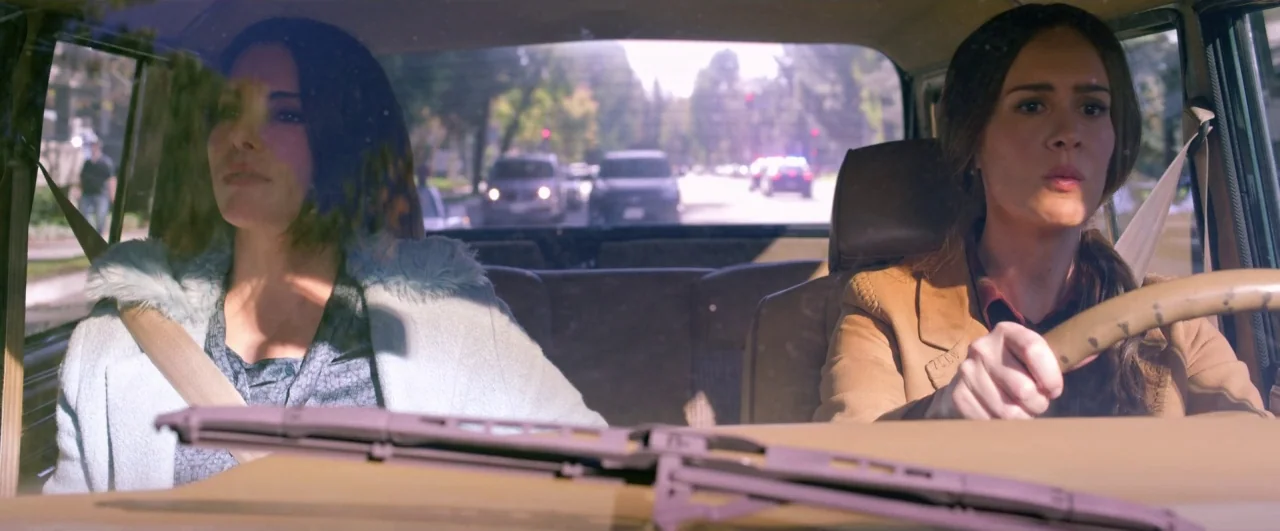Bird Box (2018) movie still: medium — Two women occupy the front seats of a car driving on a suburban road. The view through the windshield shows a…