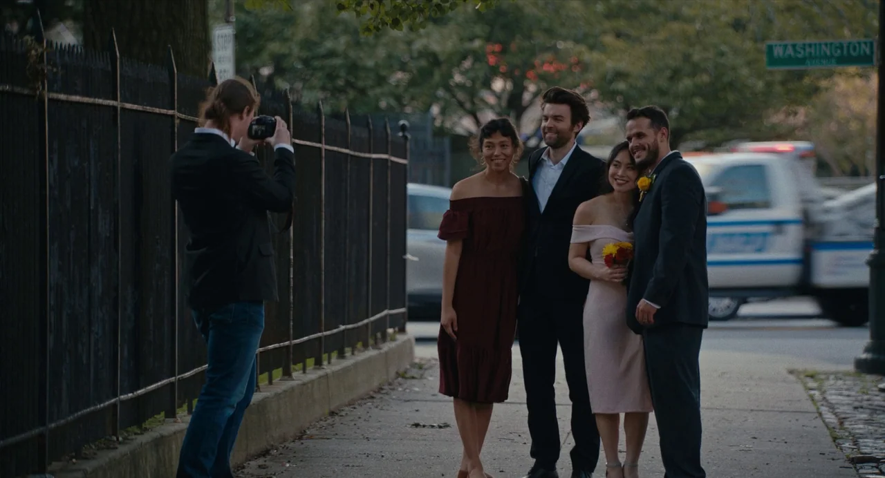Lingua Franca (2019) movie still: medium — A sidewalk next to a black wrought-iron fence on a city street. A group of four people poses for a…