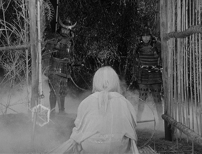 Throne of Blood (1957) movie still: fullbody — A misty outdoor space is enclosed by a tall vertical bamboo fence. A person with white hair sits…