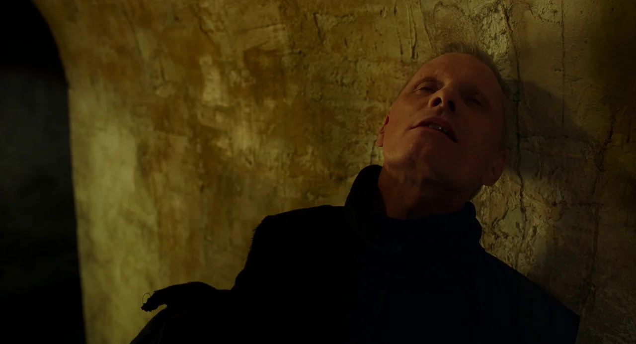 Crimes of the Future (2022) movie still: medium — An older man leans against a dimly lit textured stone wall in a vault. He rests his head against…