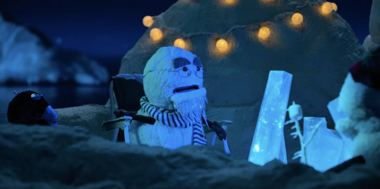 31 Minutes: One Hot Christmas (2025) movie still: medium — A puppet snowman sits in a folding chair before a constructed ice igloo structure. Warm…