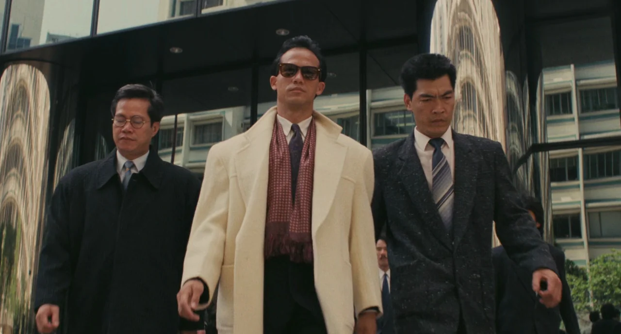 A Better Tomorrow (1986) movie still: fullbody — Three men walk in a line toward the camera past a dark reflective building. The architecture…