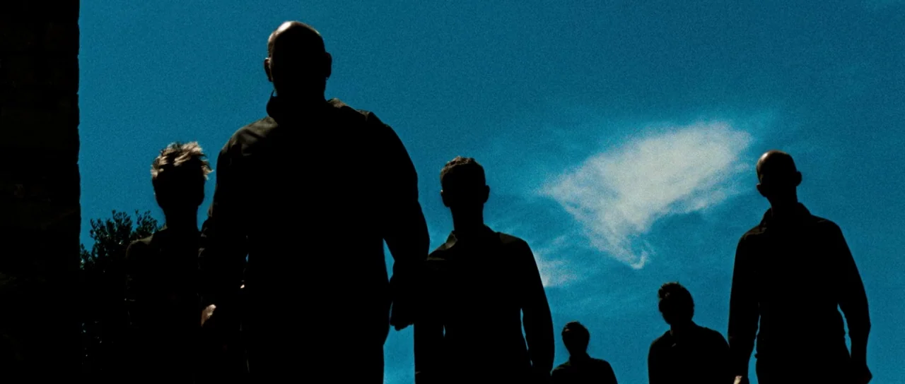 Let The Corpses Tan (2017) movie still: fullbody — A group of figures stands against a bright, clear blue sky background. The men walk toward the…