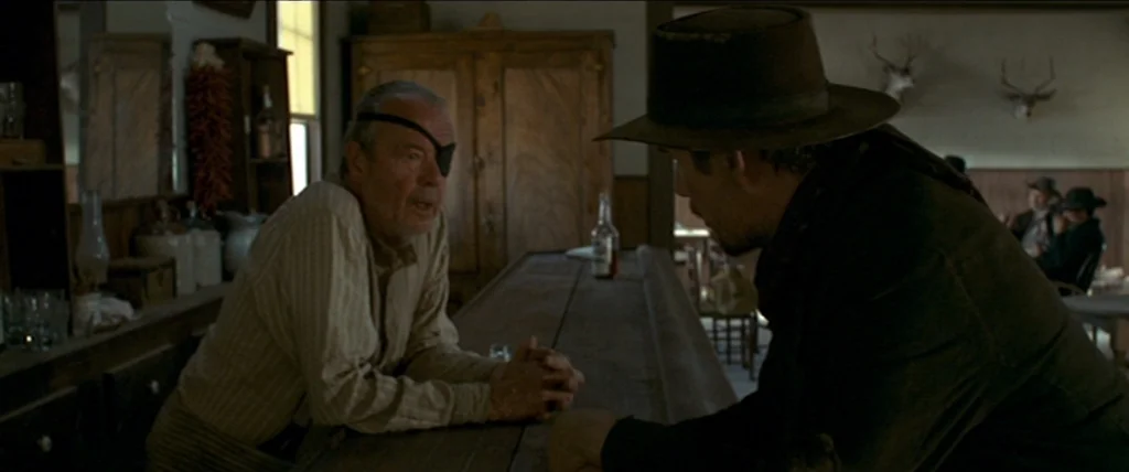 In A Valley of Violence (2016) movie still: medium — A wooden saloon interior with taxidermy deer heads mounted on the wall. Two men stand at a…