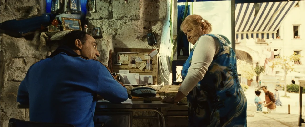 Reality (2023) movie still: medium — A rustic shop interior with exposed stone walls leads to a sunlit town plaza. A man in a blue jacket sits at a…