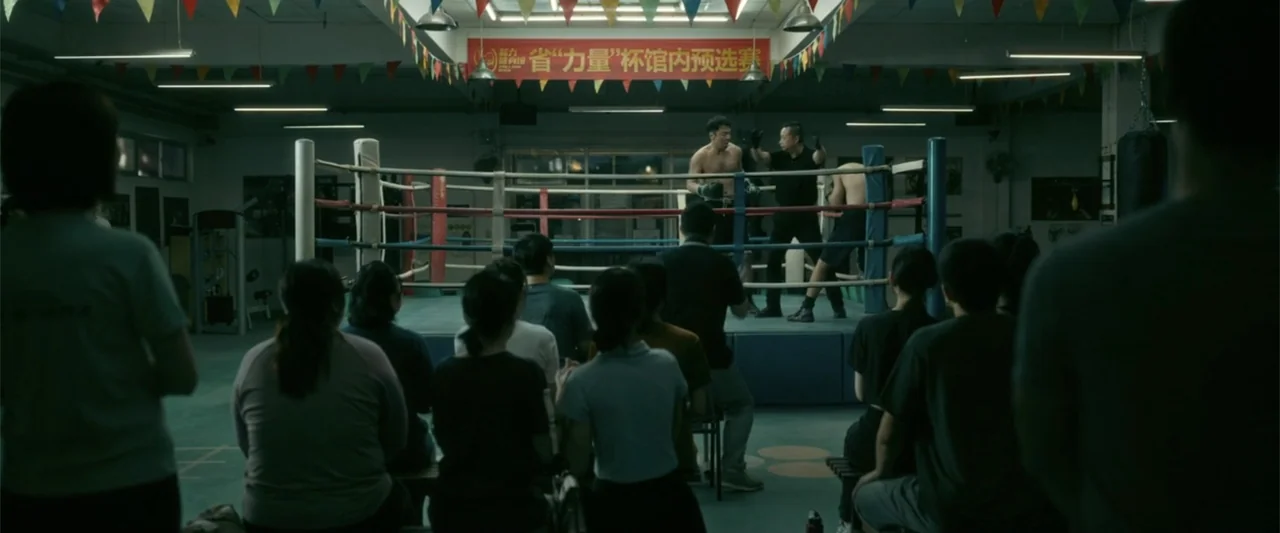 Yolo (2024) movie still: wide — A boxing gym interior features a raised ring centered under festive colorful banners. A group of spectators sits on…