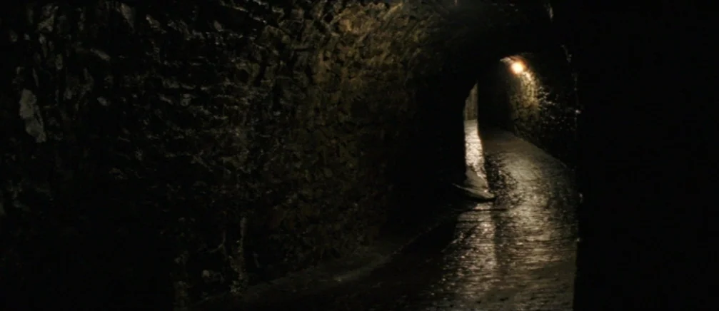Innocence (2005) movie still: establishing — A dark, narrow stone tunnel features uneven walls and a wet, slick path. A single dim light source…