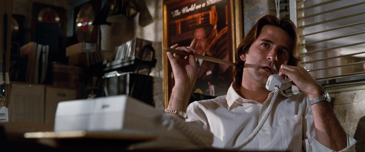 Empire Records (1995) movie still: medium — A cluttered 1990s office space features stacked record boxes and framed industry awards on walls. A man…