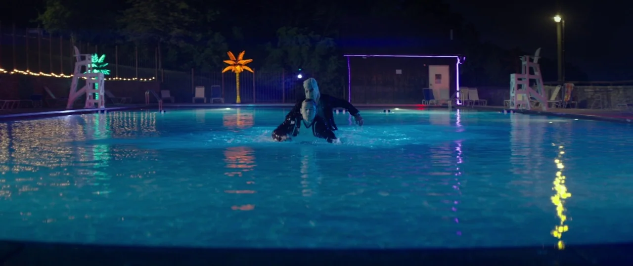 The Strangers: Prey at Night (2018) movie still: wide — A dark outdoor pool area is illuminated by neon palm trees and underwater lighting. Two men…