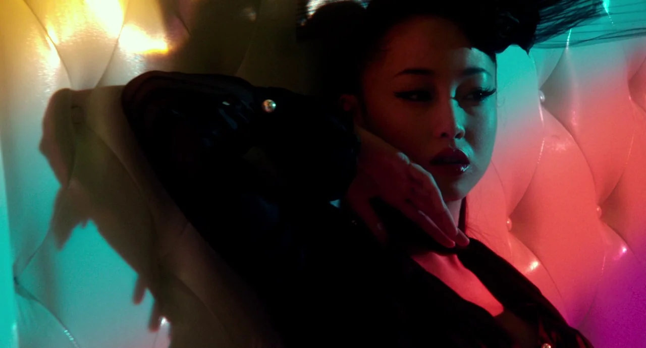 Helter Skelter (2012) movie still: closeup — A woman leans against a white button-tufted leather wall inside a neon-lit booth. She rests with her…