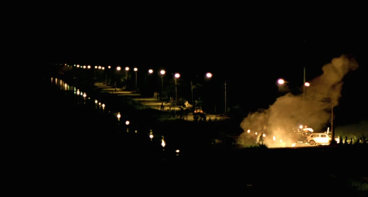 City of God (2002) movie still: wide — A dark, empty road stretches into the distance under a sequence of yellow streetlamps. A white car rests on…