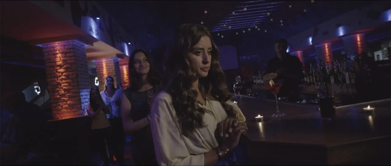 All She Wrote (2018) movie still: medium — A dimly lit lounge or bar area features exposed brick columns and wooden counters. A woman in a…