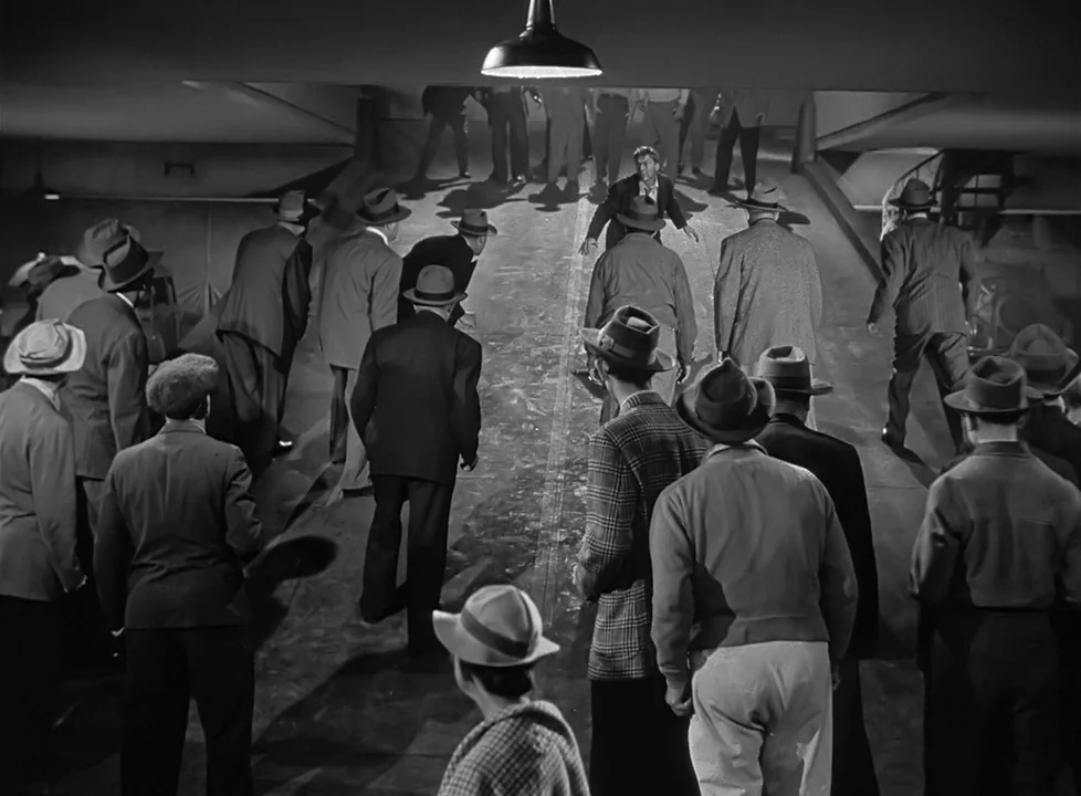 M (1951) (1951) movie still: wide — A large group of men in fedoras and suits stands in a concrete underpass. They are gathered around a central…