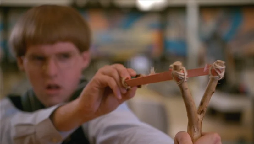 Mystery Team (2009) movie still: closeup — A man stands inside a bowling alley with a blurred mural visible in back. He grips a wooden slingshot,…