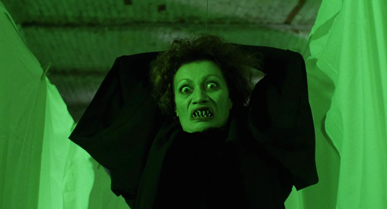The Spider Labyrinth (1988) movie still: medium — A dim area with white fabric hanging in the blurred background. A woman with prosthetic teeth…