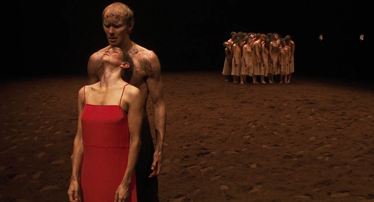 Pina (2011) movie still: fullbody — A dark stage covered in loose brown dirt features a scattered group of performers. A man and woman stand in the…