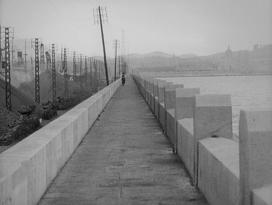 Coeur Fidele (1923) movie still: wide — A long concrete breakwater extends into the distance beside a calm body of water. A lone female figure walks…