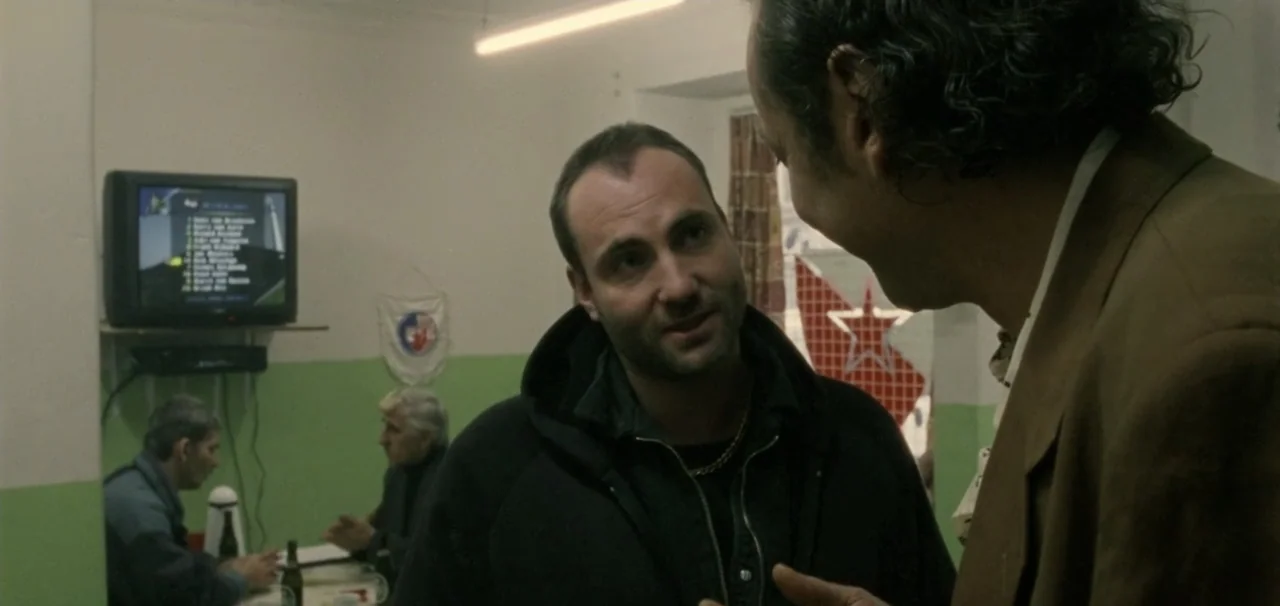 Pusher (1996) movie still: medium — A plain room features light green walls and a television mounted on the wall. A man in a dark hoodie stands in…