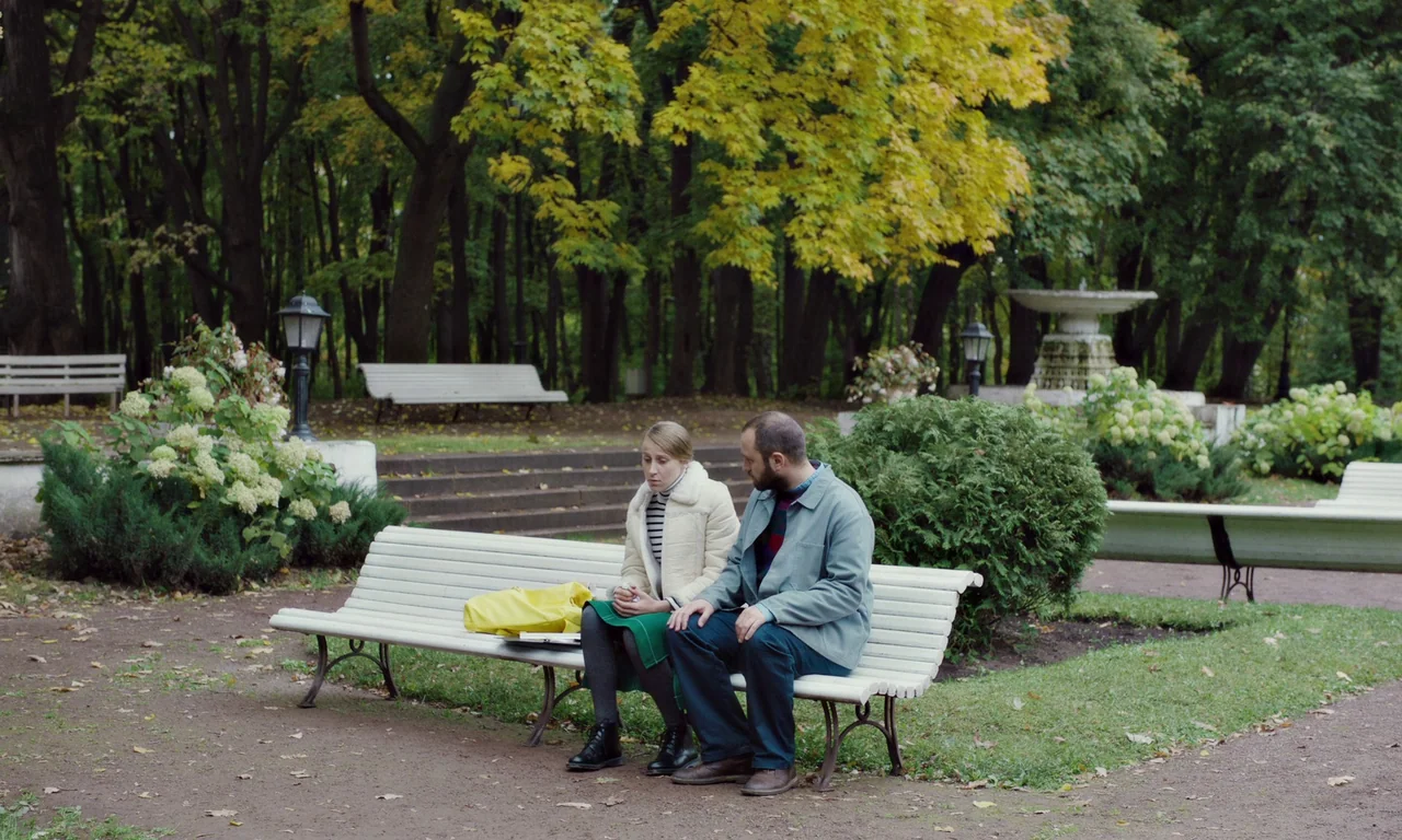 Bloodsuckers (2021) movie still: fullbody — A public park with tall trees, white wooden benches, and a fountain in back. A man and a woman sit on a…