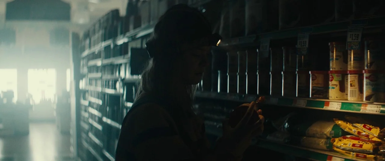 I Think We’re Alone Now (2018) movie still: medium — A dimly lit commercial aisle is filled with product shelves and thick atmospheric haze. A woman…