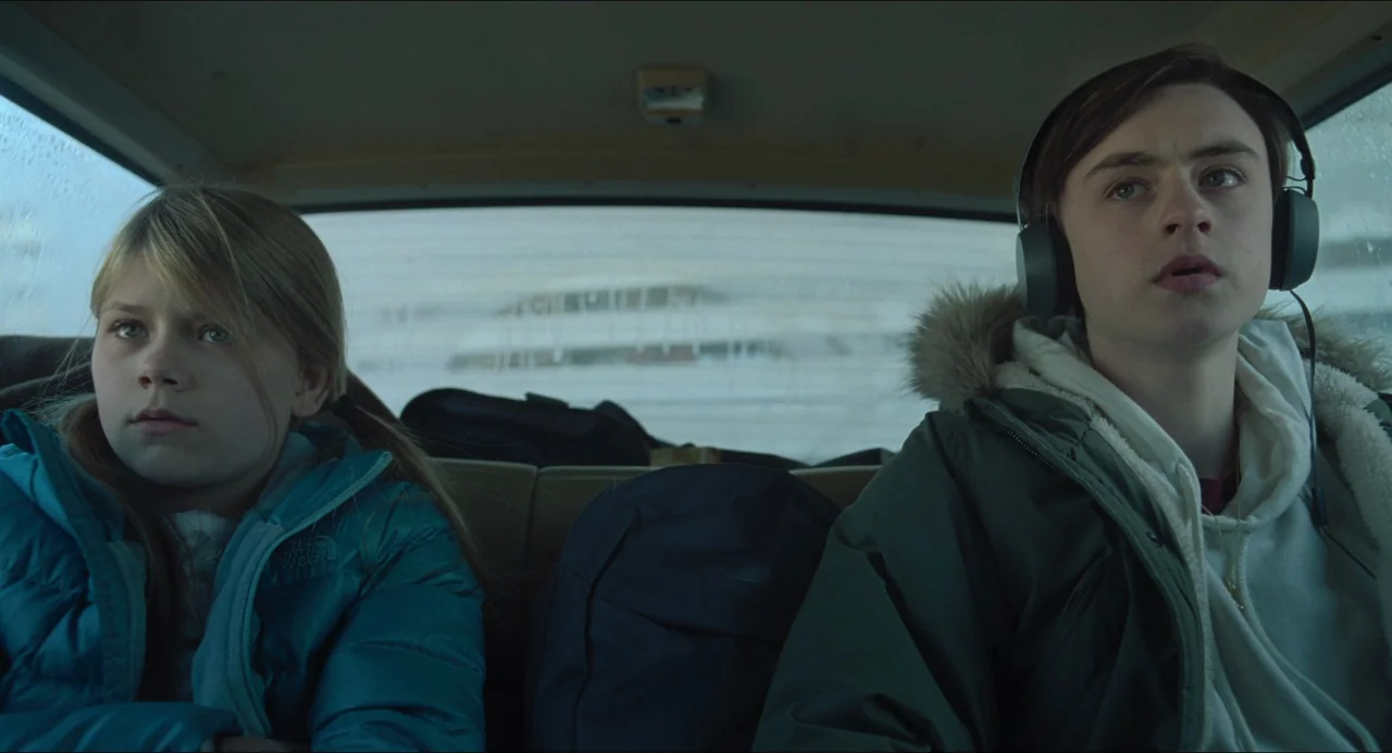 The Lodge (2019) movie still: medium — Two children sit in the backseat of a car on an overcast day. They look forward while wearing winter jackets,…