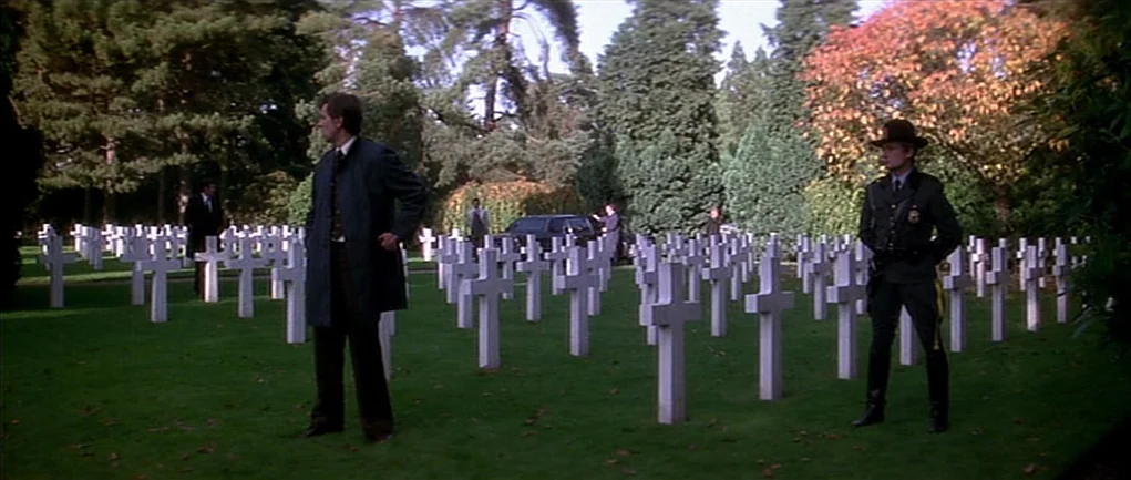 The Omen (1976) movie still: wide — A well-maintained cemetery features numerous white Latin cross grave markers on a green lawn. A man in a dark…