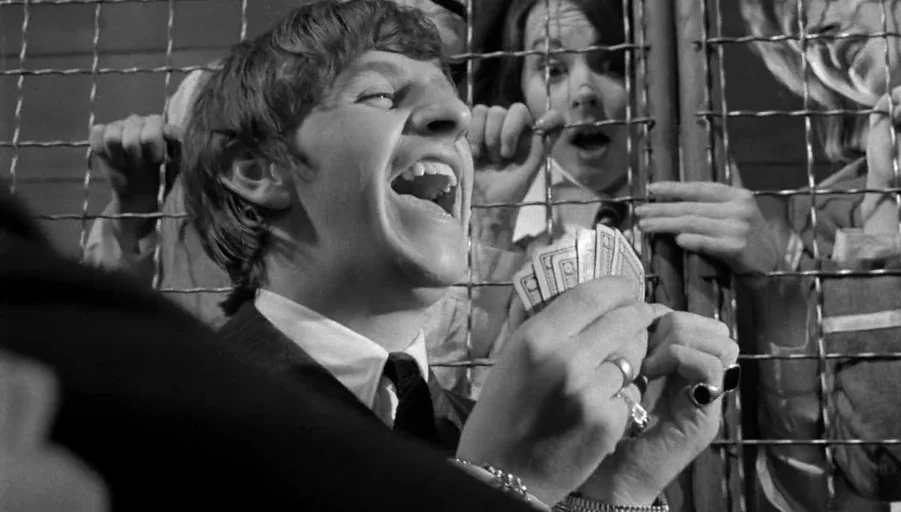 A Hard Days Night (1964) movie still: medium — A man in a suit plays cards behind a metal luggage rack barrier. A second person is visible behind…