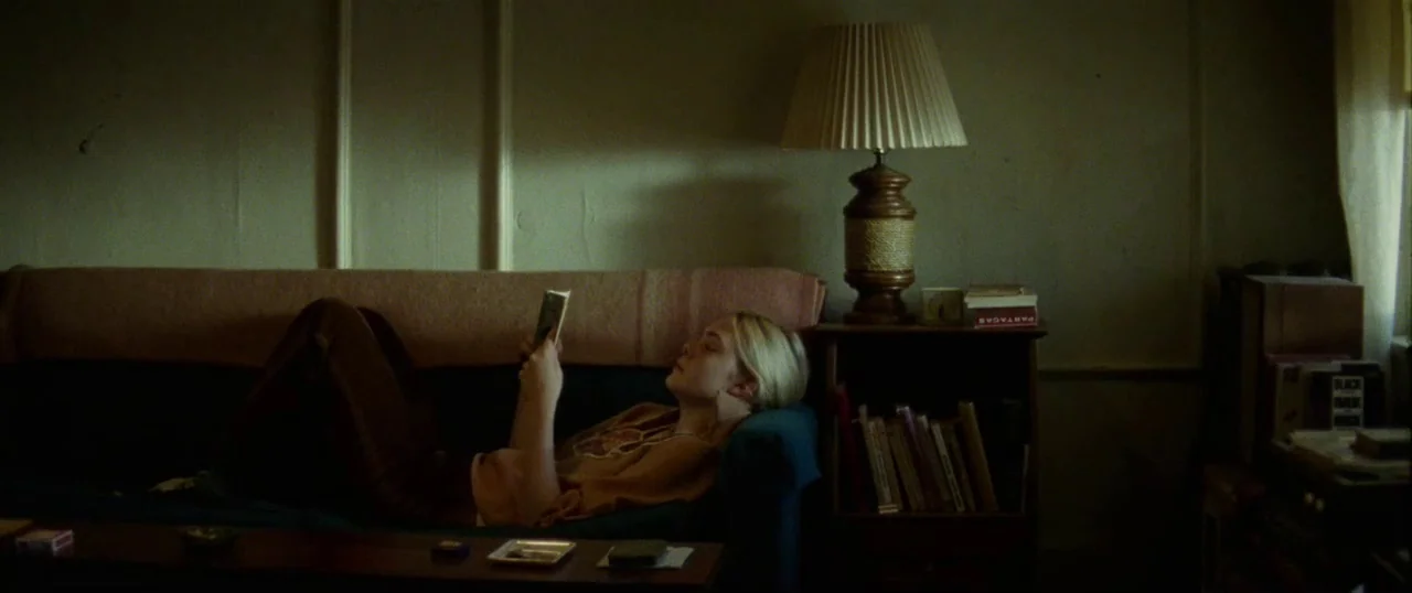 Low Down (2014) movie still: medium — A dimly lit room features a worn sofa against a paneled wall. A young woman reclines on the couch while…