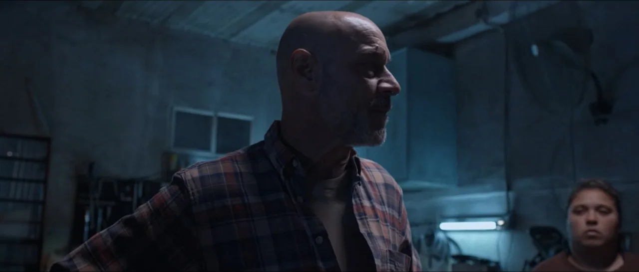 Gunman (2025) movie still: medium — A dimly lit industrial workshop with concrete walls and visible electrical equipment. A bald middle-aged man…