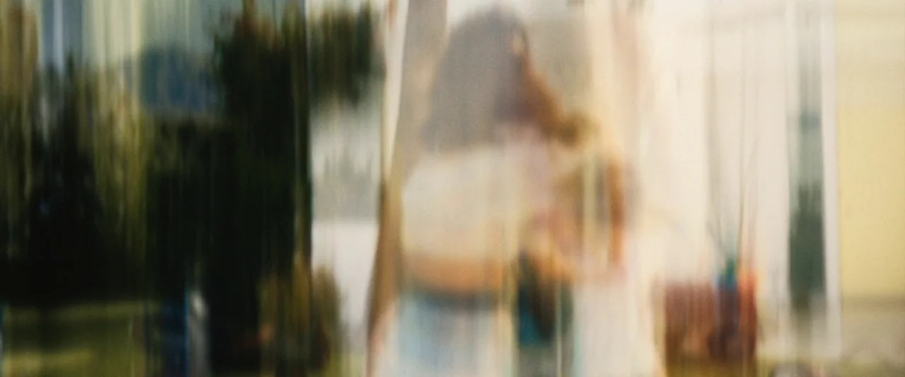 Don’t Worry Darling (2022) movie still: fullbody — An outdoor space featuring trees and light-colored structures in the distance. Two figures stand…