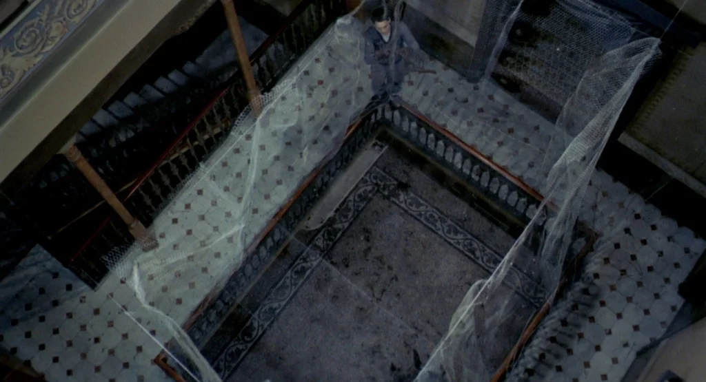 In a Glass Cage (1986) movie still: wide — An abandoned grand staircase with ornate railings is draped in dusty, translucent white netting. A lone…