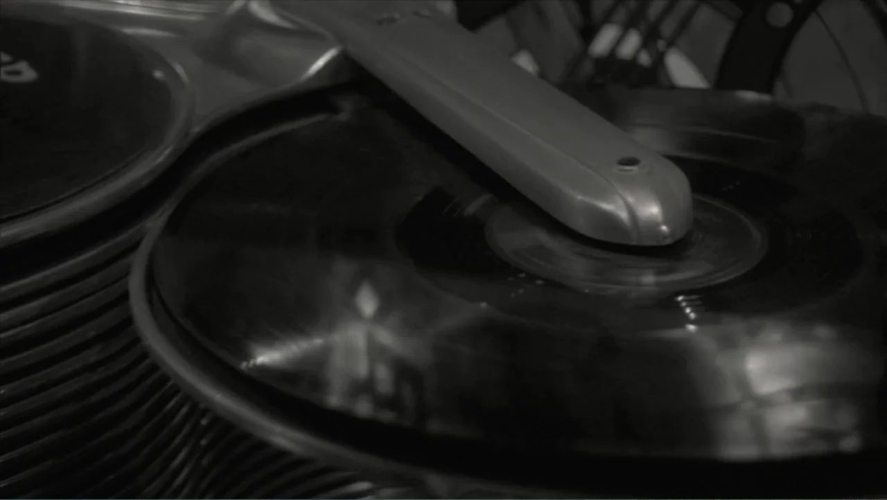The Winding Stream (2014) movie still: closeup — A close-up view shows a black vinyl record on a rotating mechanical turntable. A metallic tonearm…