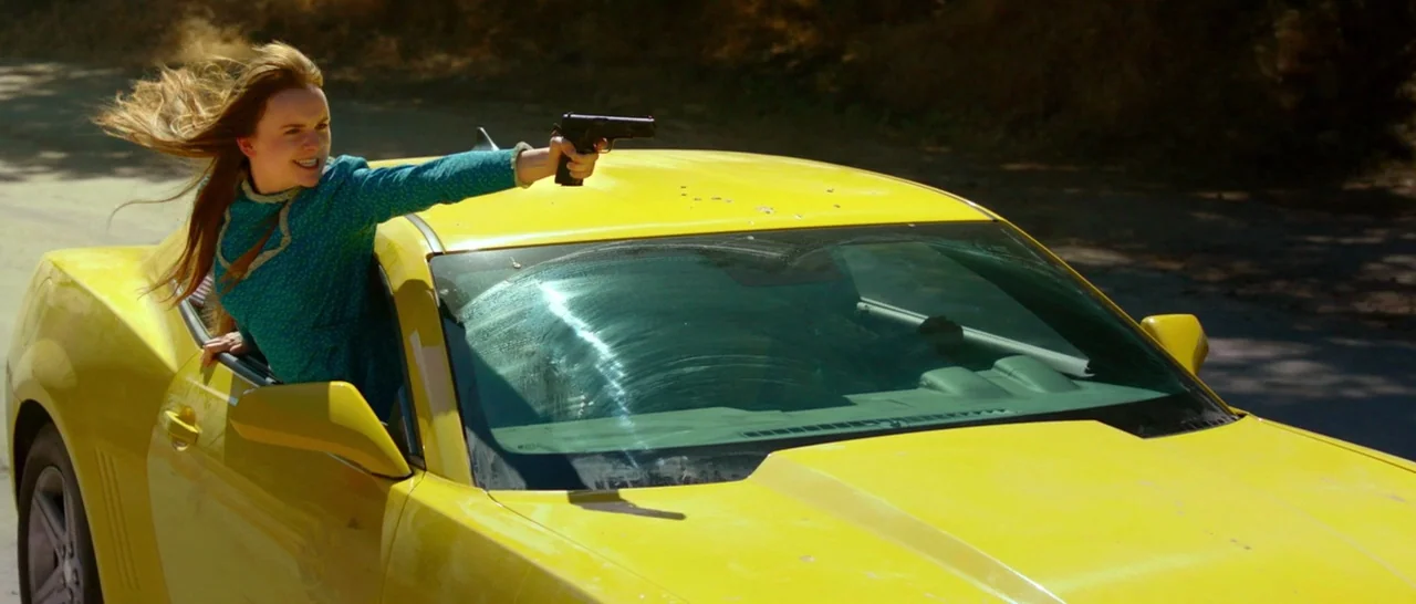 God Bless America (2011) movie still: medium — A bright yellow sports car moves along a paved road with blurred foliage behind. A young woman with…