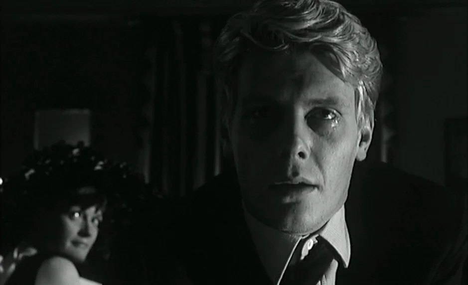 The Servant (1963) movie still: closeup — A dimly lit interior space features a man in the foreground and a woman. The man stares ahead with a tear…