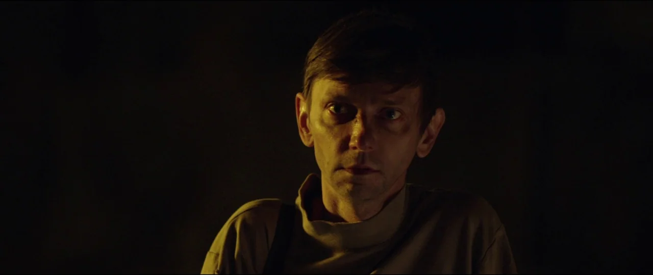 Buster’s Mal Heart (2016) movie still: closeup — A man stands in an undeterminable dark interior space. He faces the camera directly while his face…