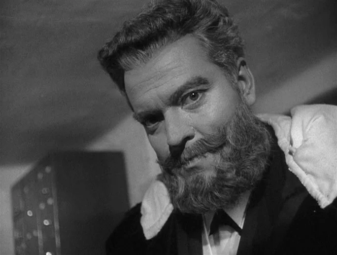 Mr. Arkadin (1955) movie still: closeup — A man with a full beard and mustache occupies the frame against a dark background. He wears a dark suit…