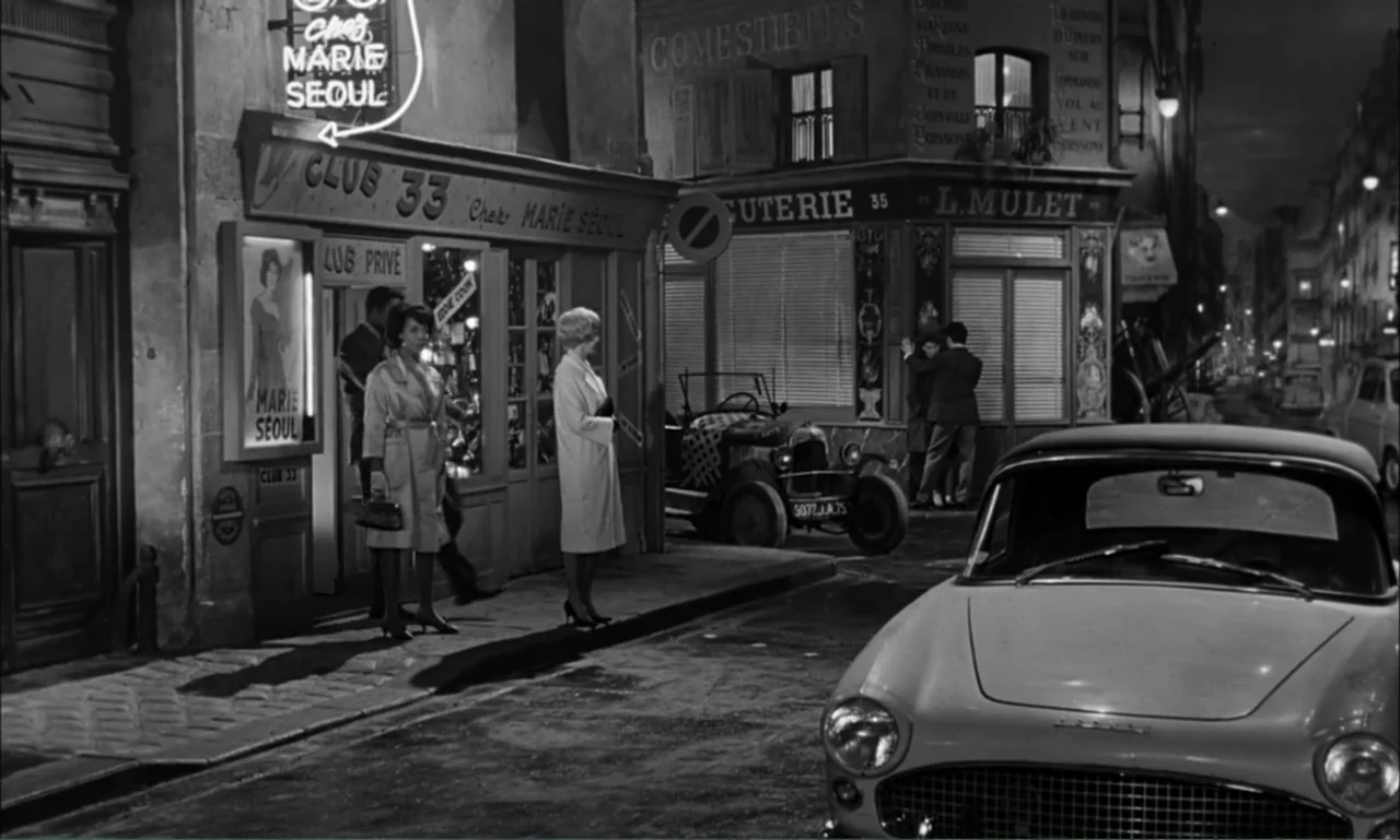 Paris Blues (1961) movie still: wide — A night street corner in a European city features storefronts and parked automobiles. Two women stand near…