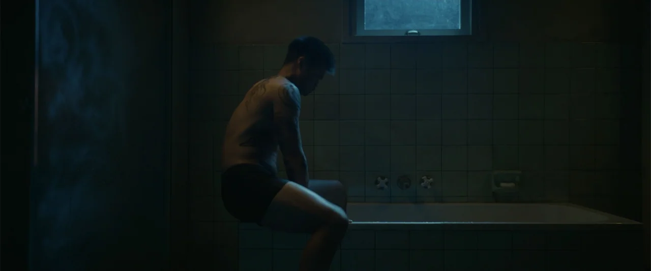 Perfect Addiction (2023) movie still: medium — A dim, teal-toned bathroom features tiled walls and a window above the bathtub. A man with tattoos…