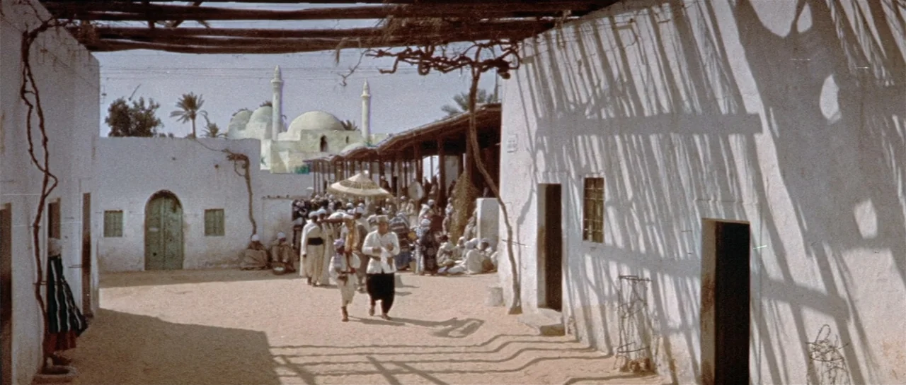 Legend of the Lost (1957) movie still: wide — A sunny outdoor courtyard features white stucco walls and a sandy ground. Several figures move through…