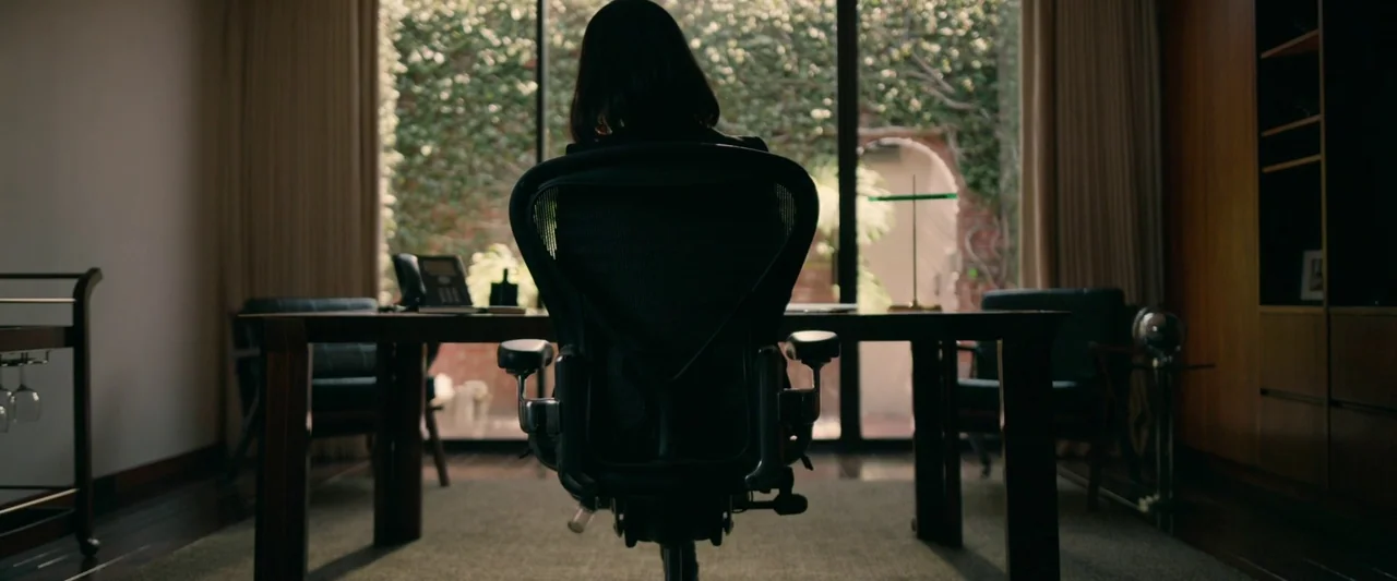 The Hunt (2020) movie still: medium — A modern room features a dark wood desk set against a bright glass window. The occupant sits in a black mesh…