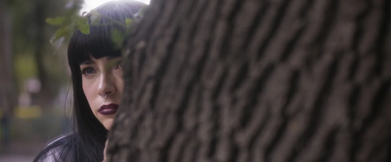 Crazy for Change (2020) movie still: closeup — An outdoor park setting features a prominent, textured tree trunk occupying the right foreground. A…