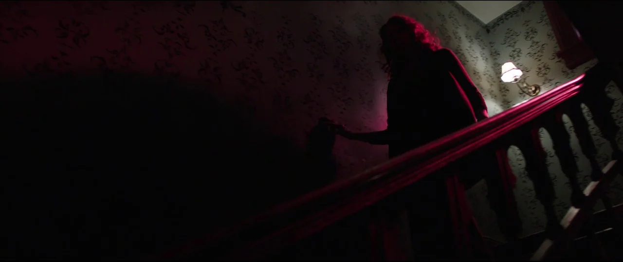 Insidious: Chapter 2 (2013) movie still: medium — A staircase with dark patterned wallpaper features a warm wall sconce and magenta light. A woman…