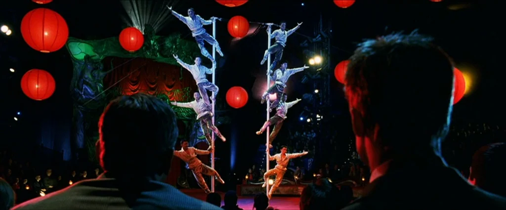 Ocean’s Eleven (2001) movie still: wide — A dark arena features performers balancing on vertical poles surrounded by glowing red lanterns. Audience…